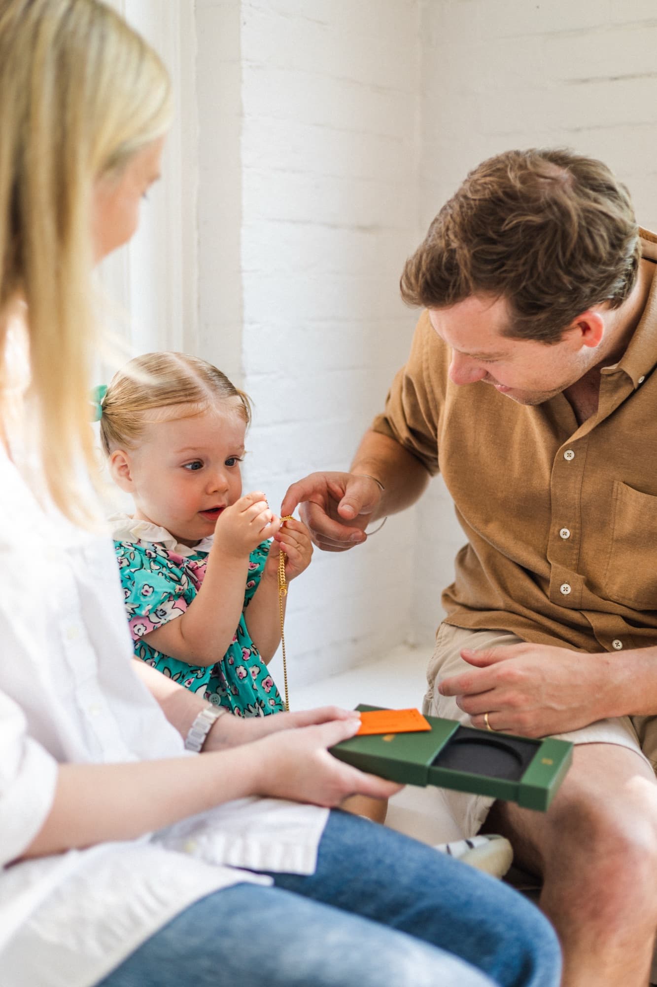 A toddler examining a gold necklace while seated between her parents